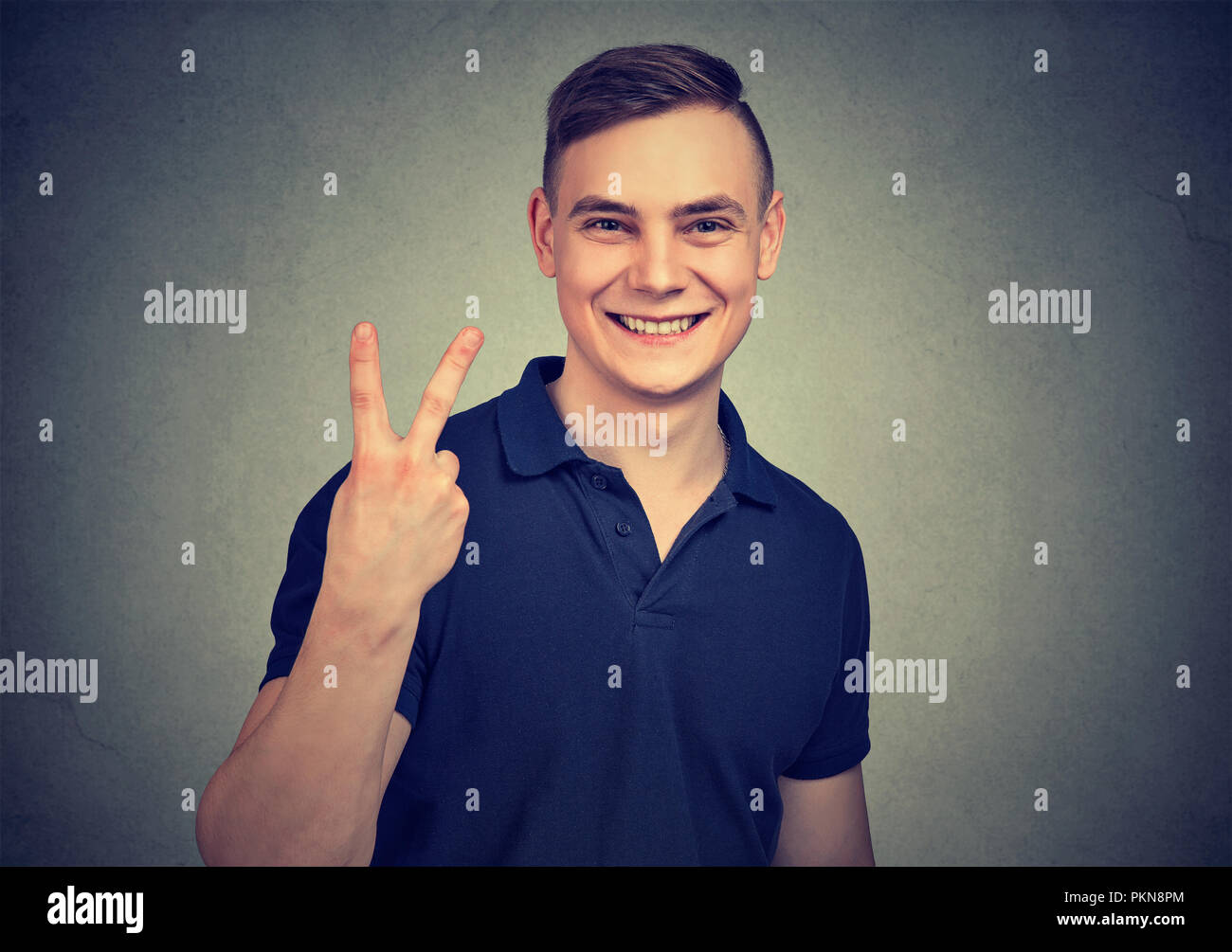 Junge legerer Mann freundlich lächelnd und mit v-Zeichen bei Kamera schaut auf grauem Hintergrund Stockfoto