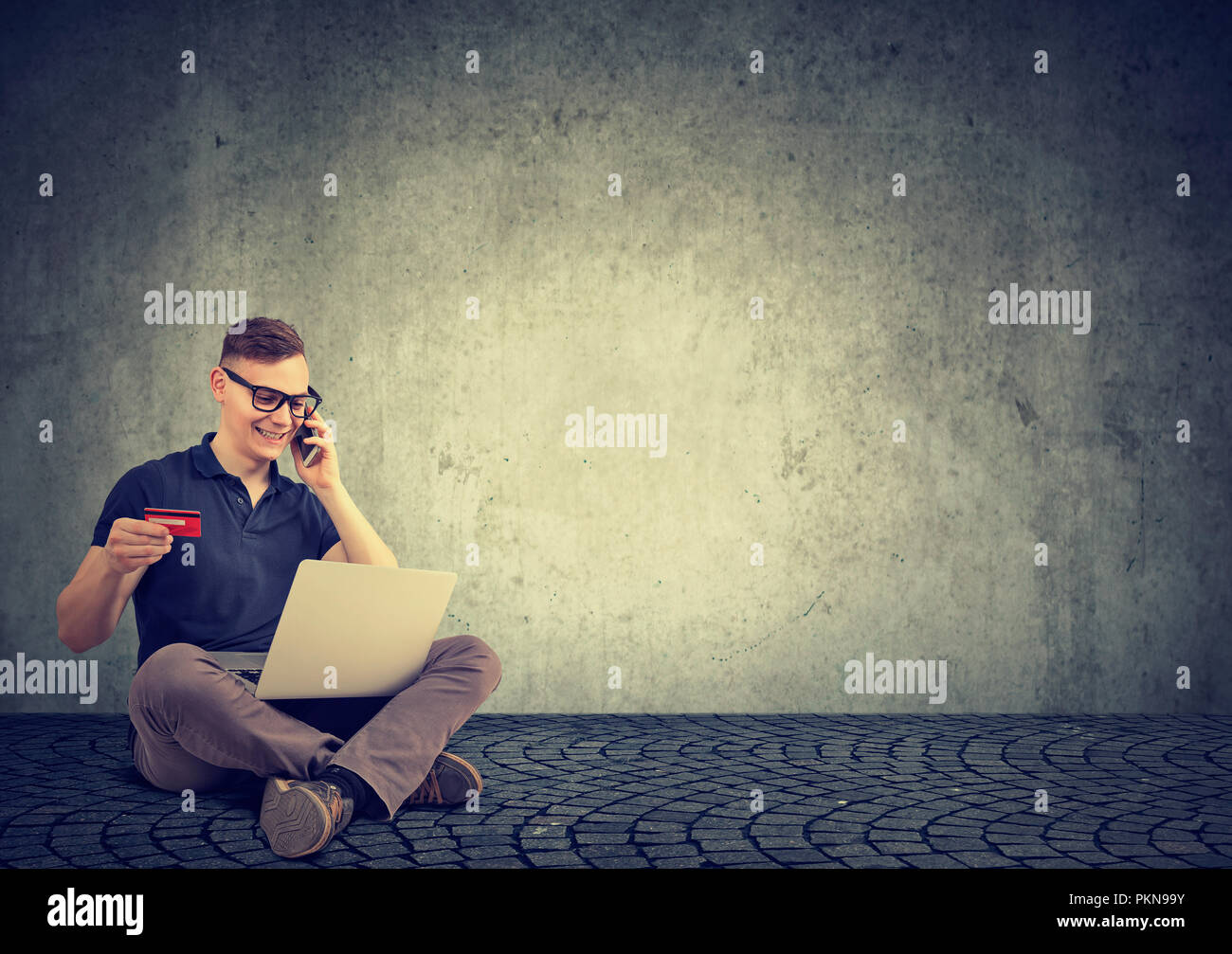 Lächelnd legerer Mann sitzen auf grauem Hintergrund mit Kreditkarte und Laptop zu kaufen, beim Sprechen am Telefon Stockfoto