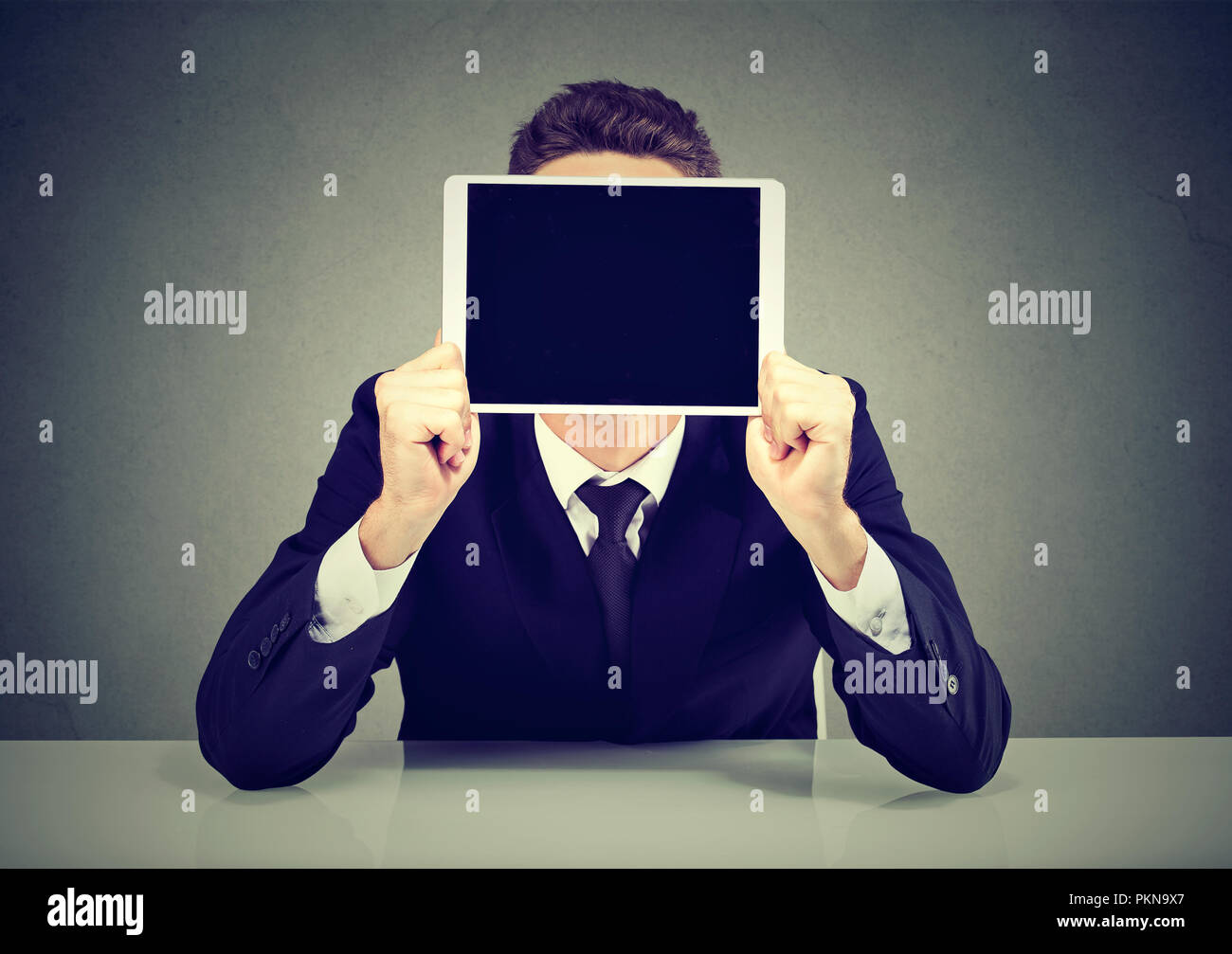 Anonyme Geschäftsmann in Anzug holding Tablet vor Gesicht sitzen auf grauem Hintergrund Stockfoto