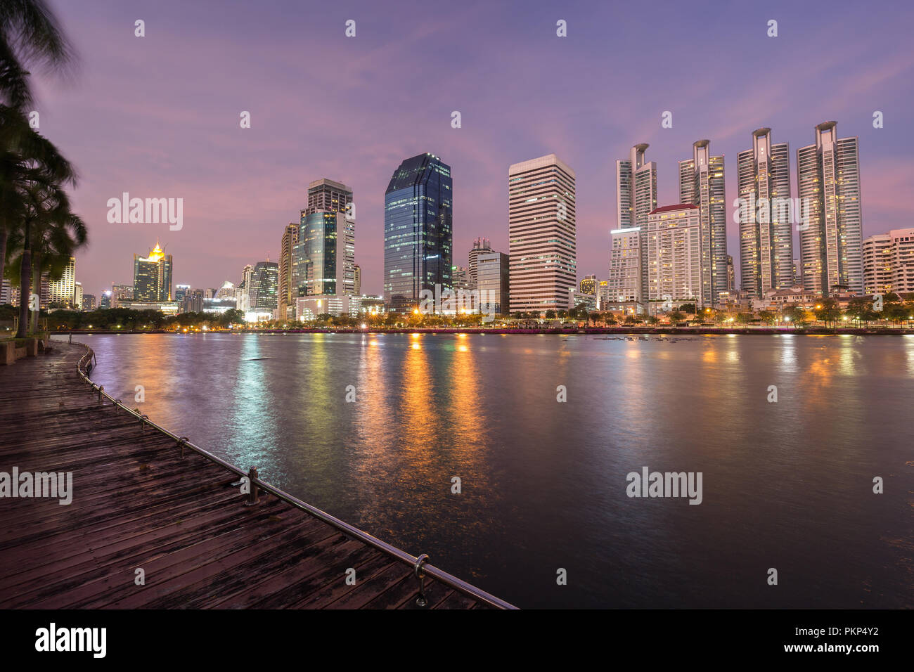 Malerischer Blick auf Holzsteg und See an der Benjakiti (benjakitti) Park und beleuchteten Wolkenkratzer in Bangkok, Thailand, bei Sonnenuntergang. Stockfoto