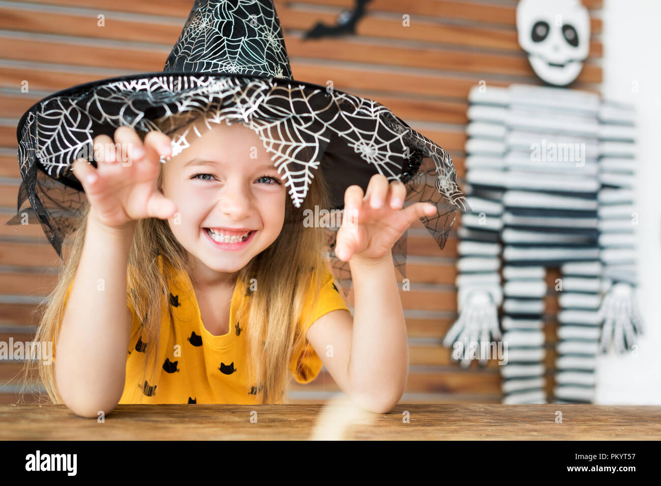 Süße kleine Mädchen mit Hexenhut hinter einem Tisch in Halloween Thema eingerichteten Wohnzimmer sitzen, an der Kamera schaut und beängstigend. Halloween Stockfoto