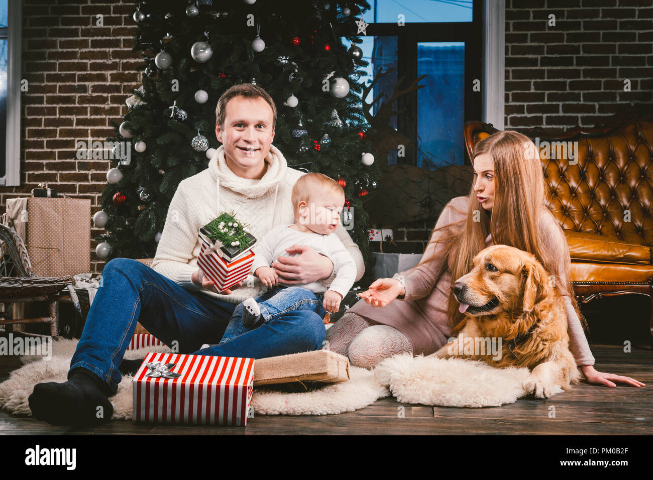 Thema Weihnachten und Neues Jahr Familie Kreis- und Haustier. Mama, Papa und Kind 1 Jahr alt kaukasische Frau sitzt auf dem Boden in der Nähe der Weihnachtsbaum mit Gi Stockfoto