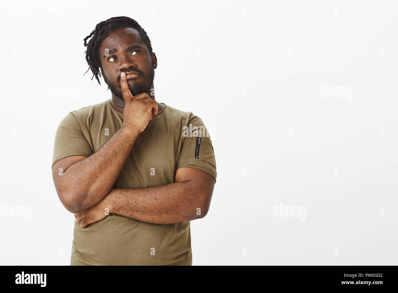 Studio shot von neugierig Dunkelhäutige gewöhnlicher Kerl mit Bart in Oliv t-shirt, und halten Sie den Finger auf der Lippe beim Denken, die Entscheidung bzw. Wahl, stehend nachdenklich über grauer Hintergrund Stockfoto