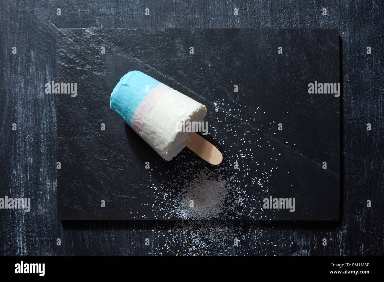 Bunte Eis auf einem Stick mit Kokosnuss crumb auf schwarzem Schiefer auf einem dunklen konkreten Hintergrund mit Kopie Raum dargestellt. Flach Stockfoto