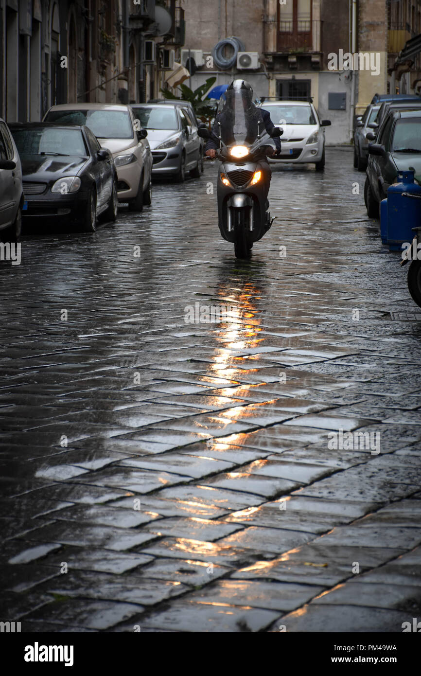 Mann, ein kleines Motorrad durch die Straßen von einer italienischen Stadt, mit dem headlinght im nassen Stein Pflaster der Straße reflektierenden Stockfoto