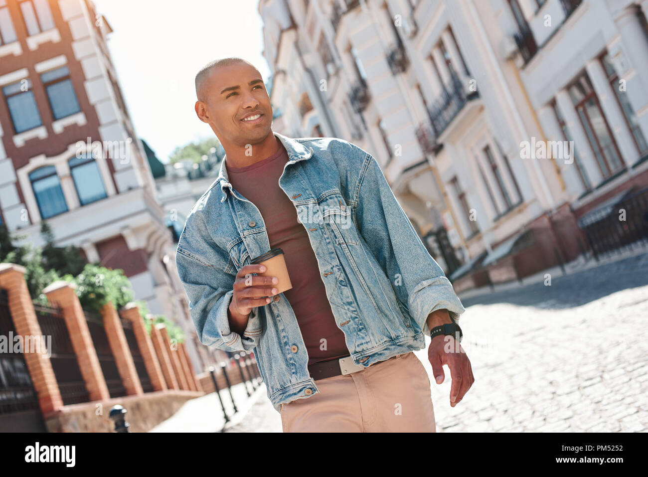 Morgen Energie. Junge Mann zu Fuß auf city street Holding Tasse trinken heißen Kaffee zur Seite schauen, lächeln fröhlicher Stockfoto