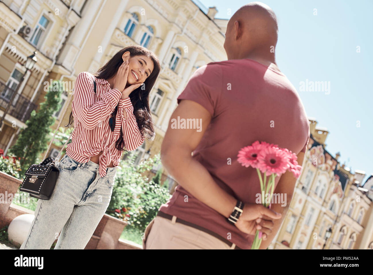 Romantische Überraschung. Junge diverse Paar stand auf der Straße der Stadt Freund holding Blumenstrauß hinter Rücken, während Freundin auf ihn schaut lächelnd heiter Stockfoto