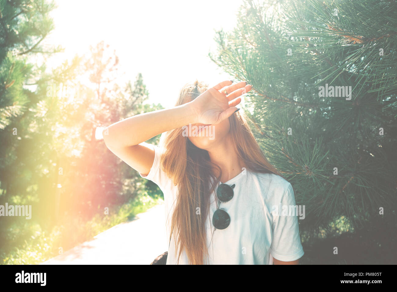 Close-up junge Frau mit schönen Gesicht verfügt über ihr Gesicht von Sonnenstrahlen mit Arm verstecken, Pinien im Hintergrund. Licht Filter verwendet. Stockfoto
