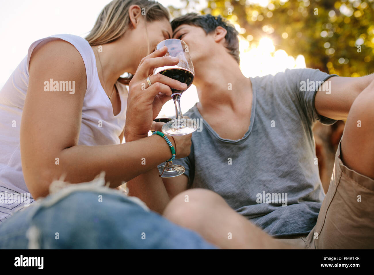 Paar Sitzen im Freien halten Glas Wein küssen einander. Paar im Park trinken Wein unter einem Baum mit Sun flare im Hintergrund sitzen. Stockfoto