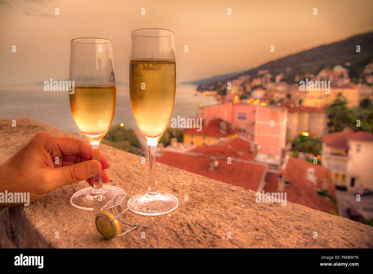 Zwei gefüllte Champagnergläser mit eines Menschen Hand ein Glas auf einem Balkon mit Blick auf das Dorf und die Adria in Kroatien Holding Stockfoto
