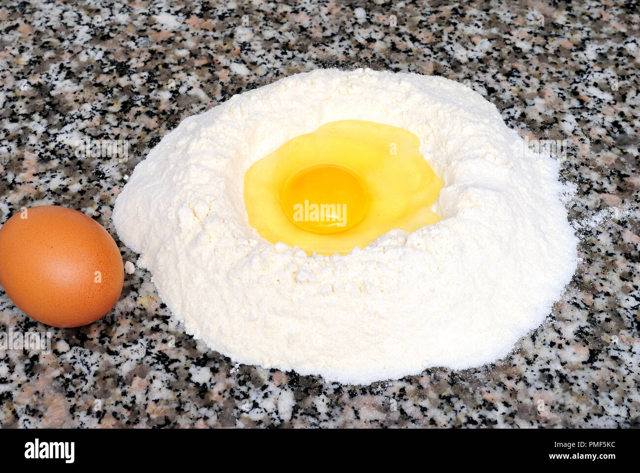 Mehl Brunnen mit einem Ei im Zentrum, neben dem ein ganzes Ei, flach Marmor Hintergrund, die zutaten Desserts und home Teig vorzubereiten Stockfoto