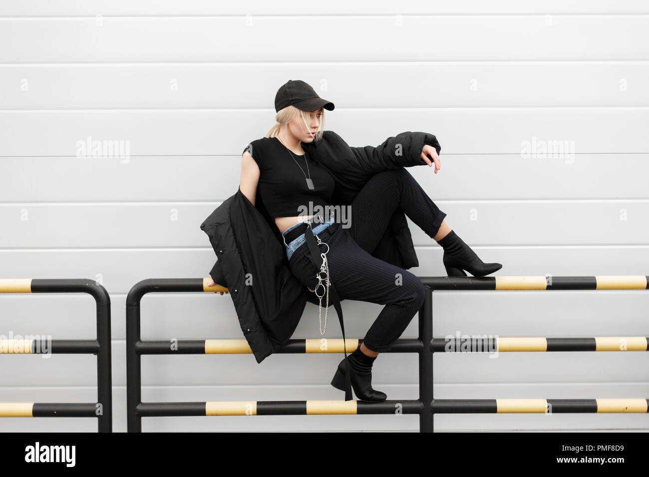 Schöne Mode Modell Frau in schwarzem stilvolle Gap und Winterjacke mit modischen Jeans - Hosen und Stiefel sitzen auf der Straße in der Nähe der graue Wand Stockfoto