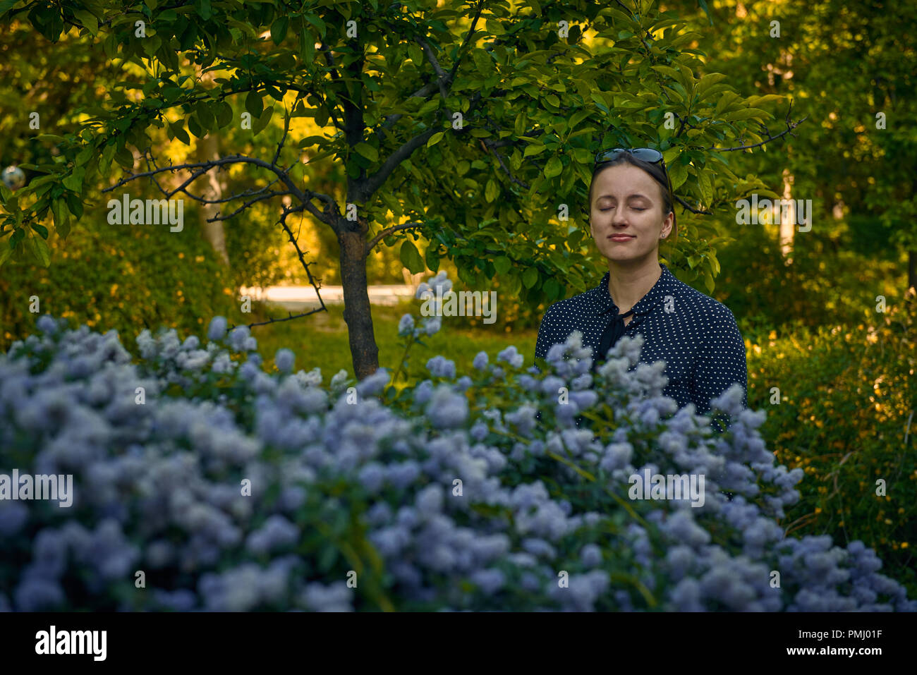 Lächelnden jungen kaukasischen Frau mit geschlossen Augen stehen in der Nähe der blühende Strauch mit blauen Blumen im öffentlichen Park Stockfoto