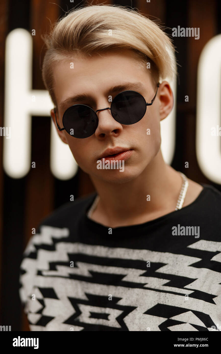 Stilvolle jungen hübschen Modell Kerl mit einer Frisur in runde Sonnenbrille in einem schwarzen Jersey in der Nähe von eine Holzwand mit Buchstaben Stockfoto