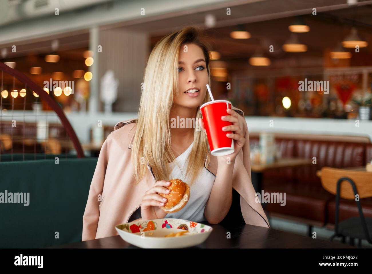 Schöne junge Modell Frau in einem rosa Mode Jacke essen fast food in einem Café, Getränke eine Cola aus einem roten Schale Stockfoto