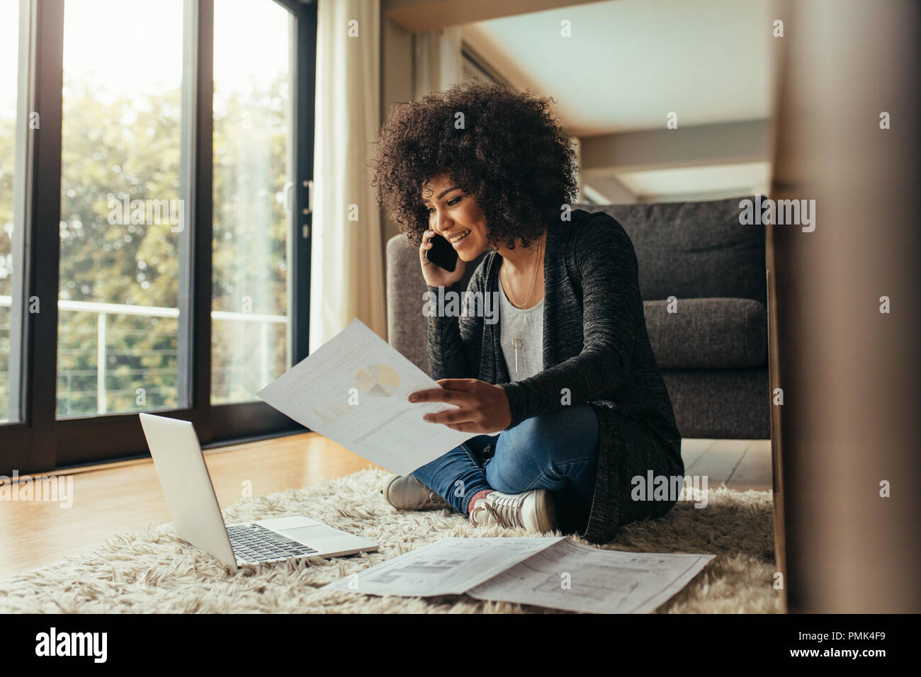 Lächelnde junge Frau sitzt auf dem Boden mit wenigen Berichte Gespräch am Handy. Afrikanische Frauen von zu Hause aus arbeiten. Stockfoto