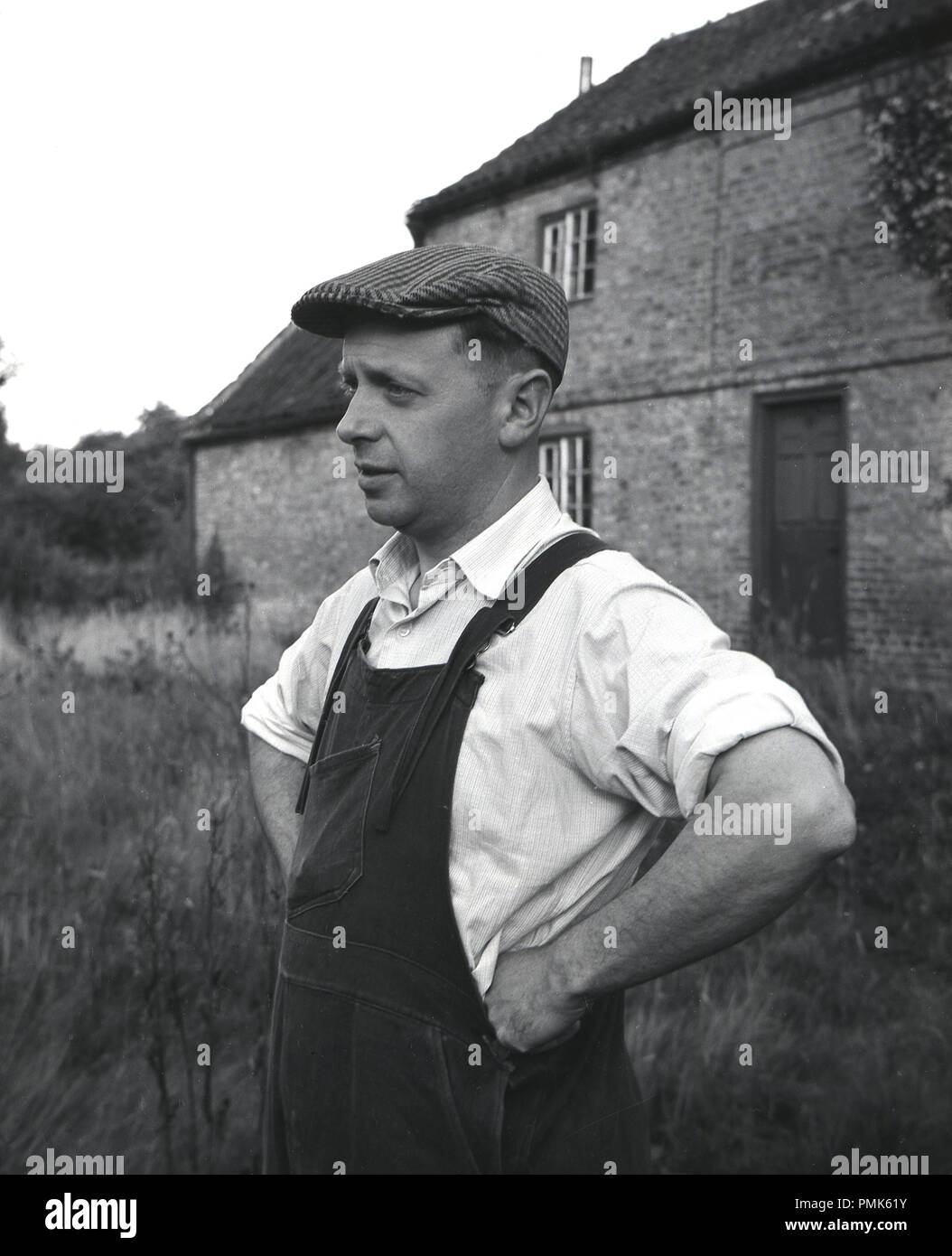 1950, historische, Porträt einer männlichen Immobilien Hausmeister oder Handwerker außerhalb ein Bauernhof, seine Arbeitskleidung, Hemd mit Kragen, dunagarees und flache Kappe tragen, England, UK. Stockfoto