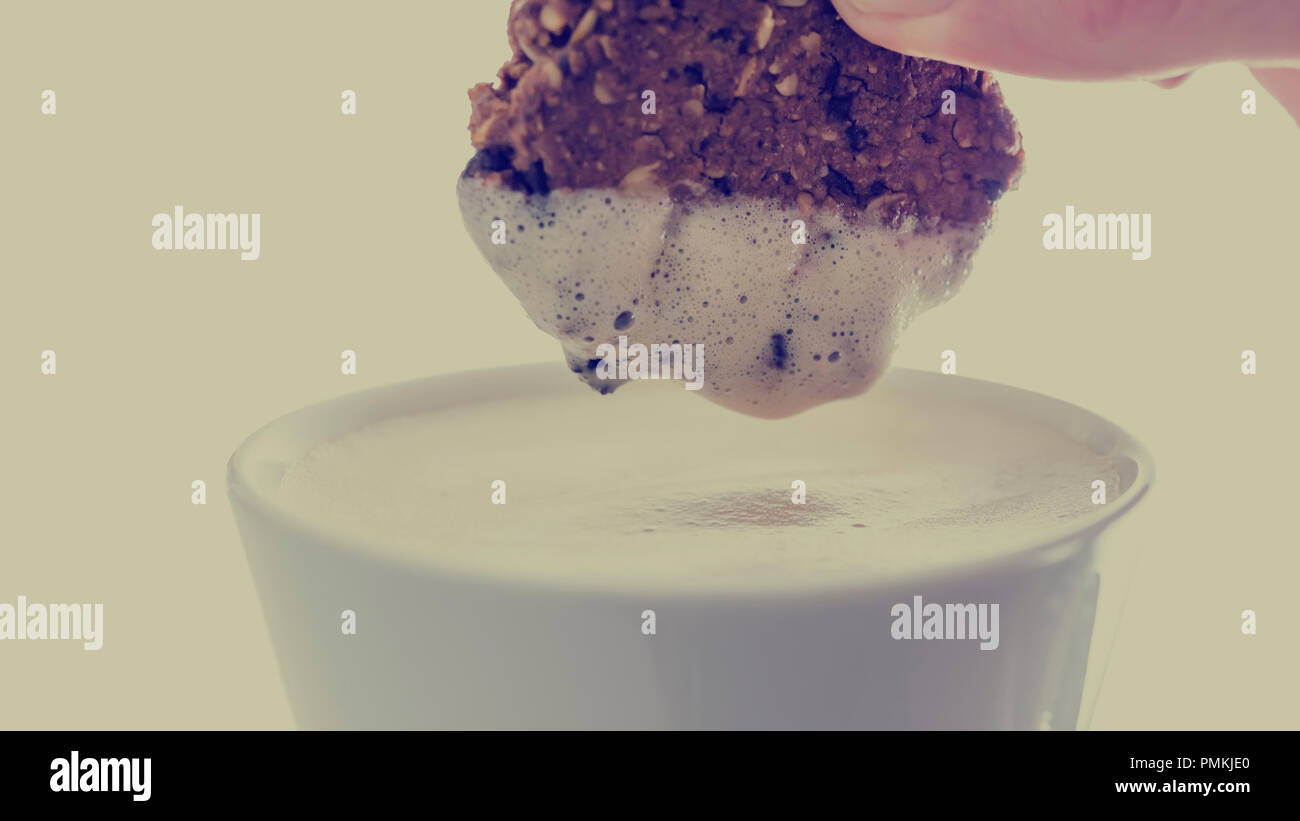 Person DUNKING einen kleinen knusprigem Keks in einer Tasse cremiger Latte oder Cappuccino mit Milchschaum die Einhaltung der Cookie in retro Farben Wirkung. Stockfoto