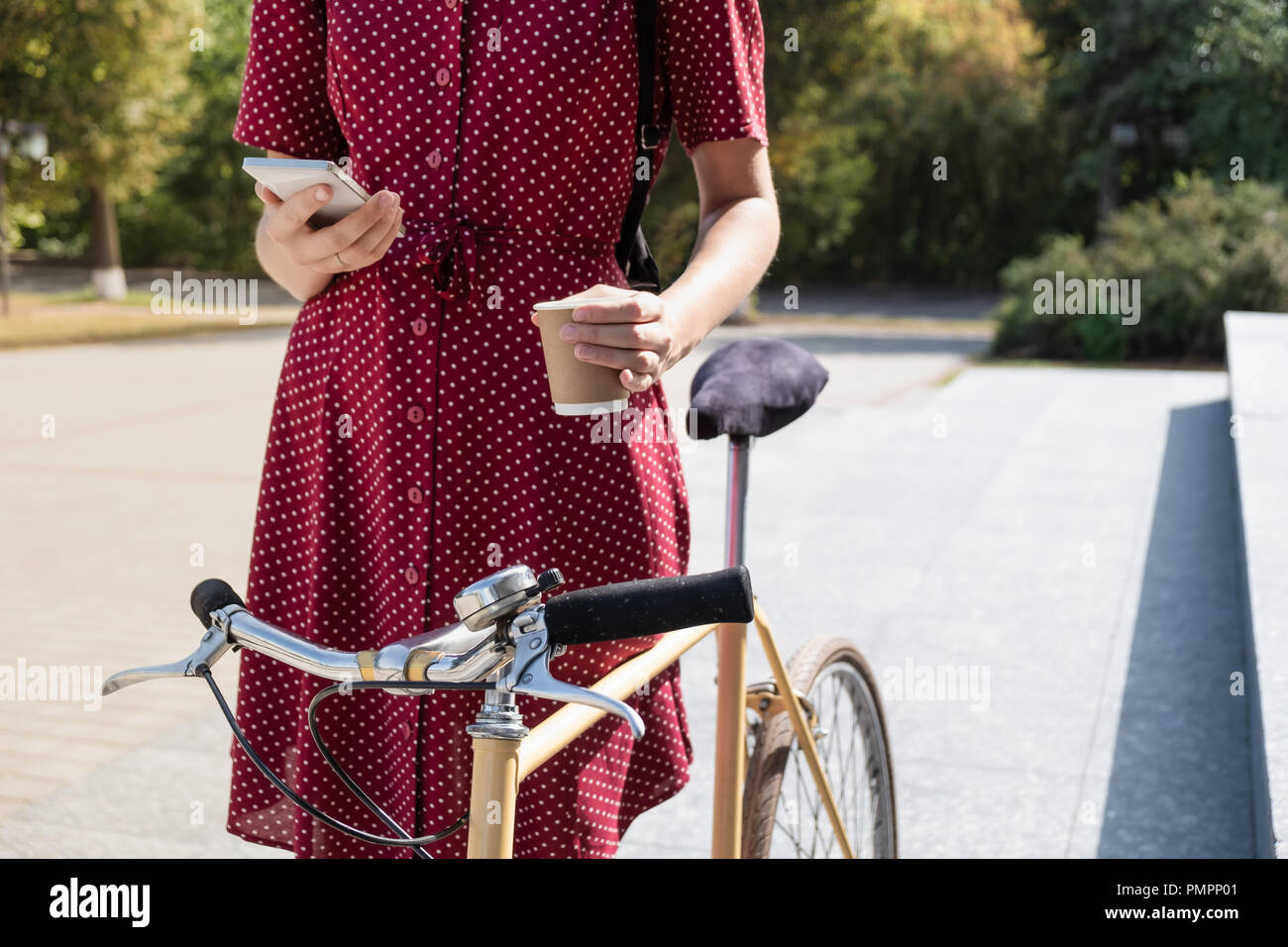 Frau in Polka Dot Kleid mit dem Fahrrad pendeln, Abrufen von E-Mail online und Kaffee. Weibliche Person mit Smartphone und Holding Tasse heißen Getränk auf Stockfoto