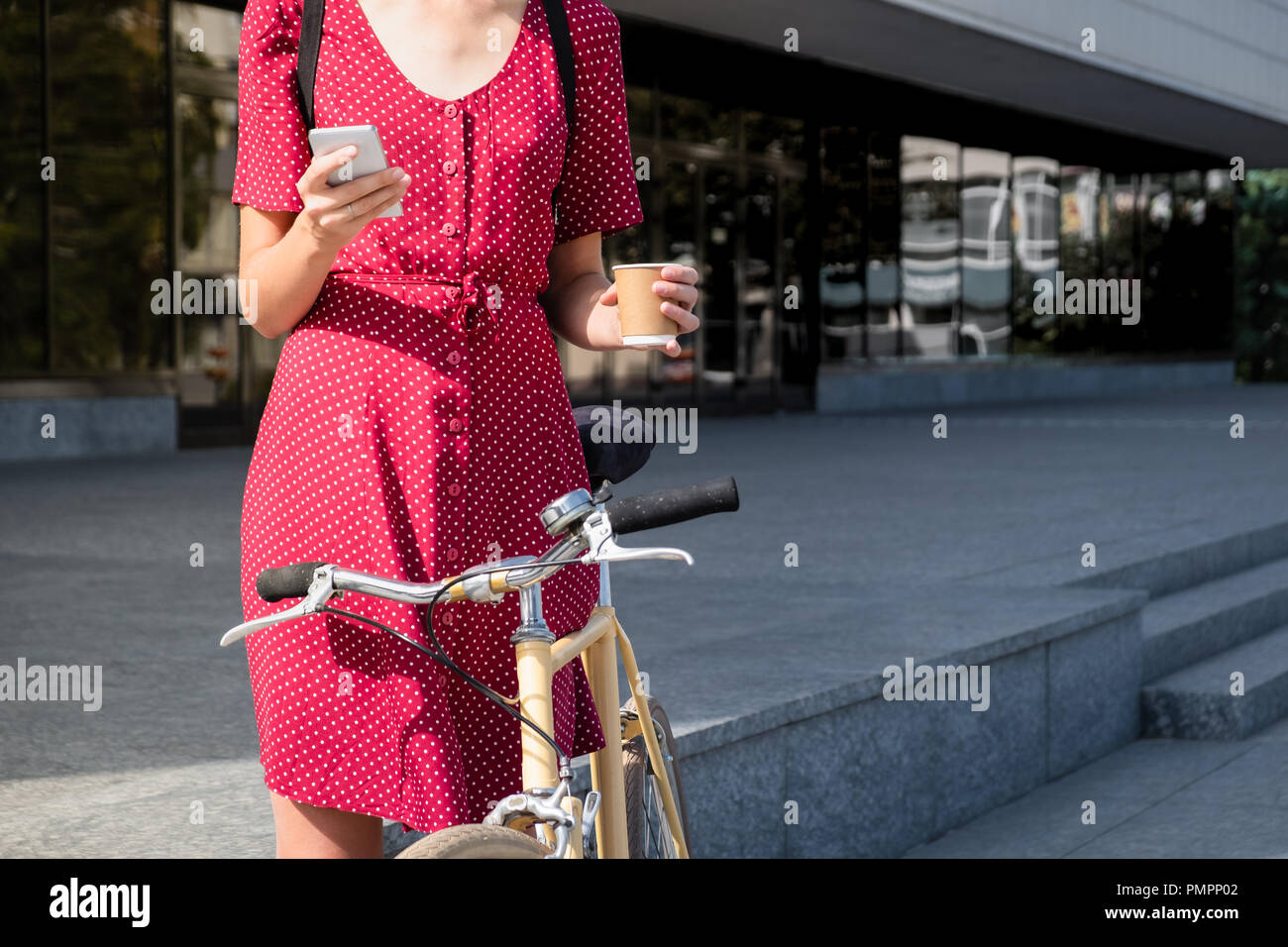 Frau in Polka Dot Kleid mit dem Fahrrad pendeln, Abrufen von E-Mail online und Kaffee. Weibliche Person mit Smartphone und Holding Tasse heißen Getränk auf Stockfoto