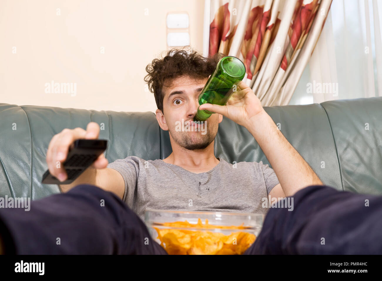Junger Mann mit der Fernbedienung und Bier und Chips auf der Couch aufgeregt Stockfoto