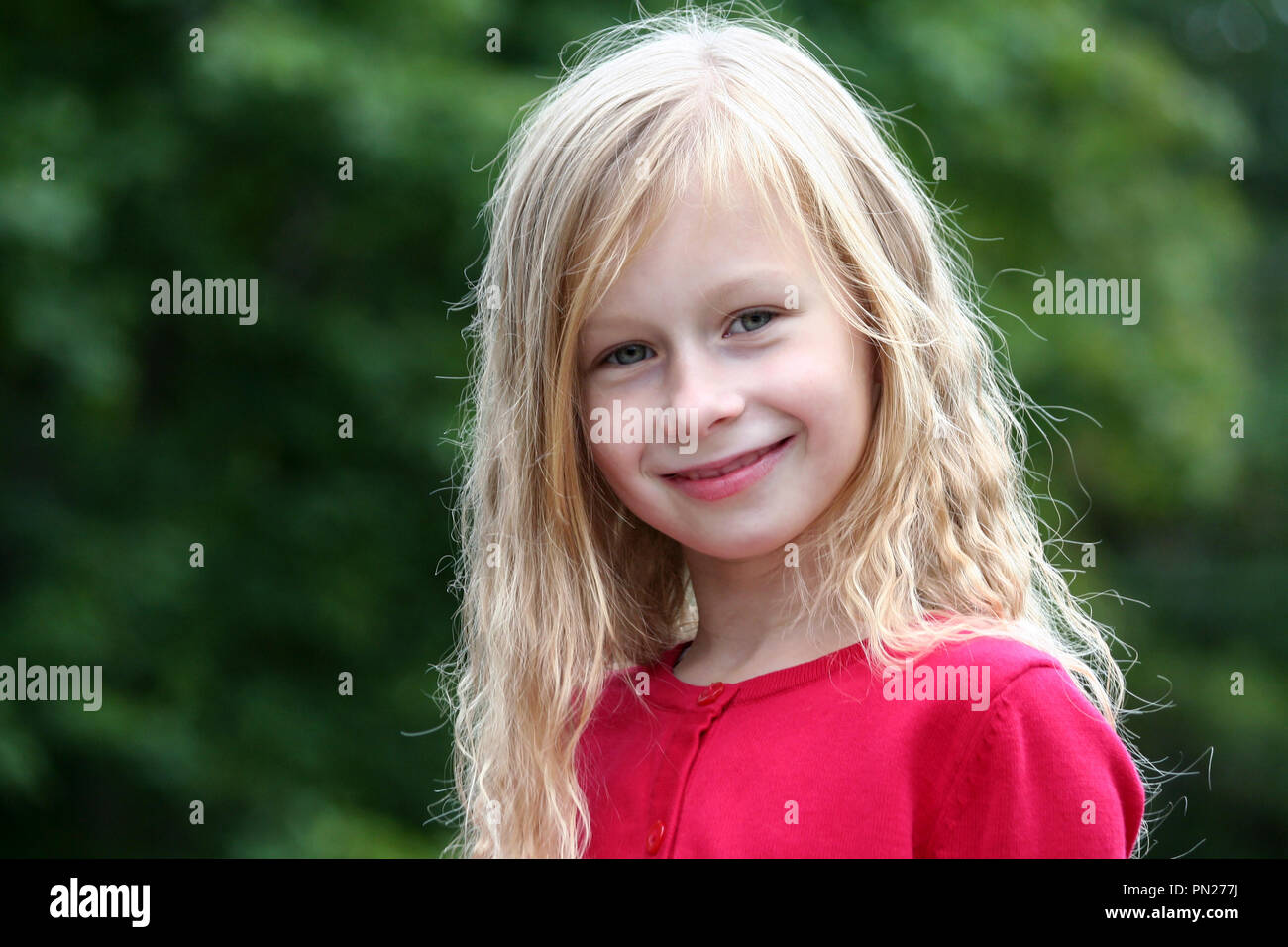 Portrait kleine Mädchen mit langen blonden Haaren in einem roten Pullover lächelnd und direkt auf die Kamera im Hintergrund, grüne Bäume, dunkles Laub, Stockfoto