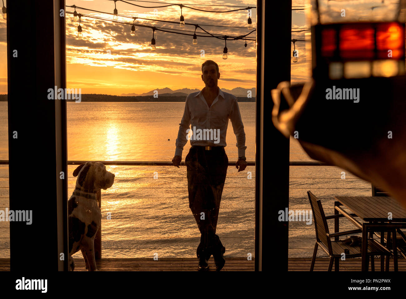 Silhouetted Mann mit Dogge Hund auf Deck in Beach home mit einem Glas Alkohol im Vordergrund bei Sonnenuntergang. Stockfoto