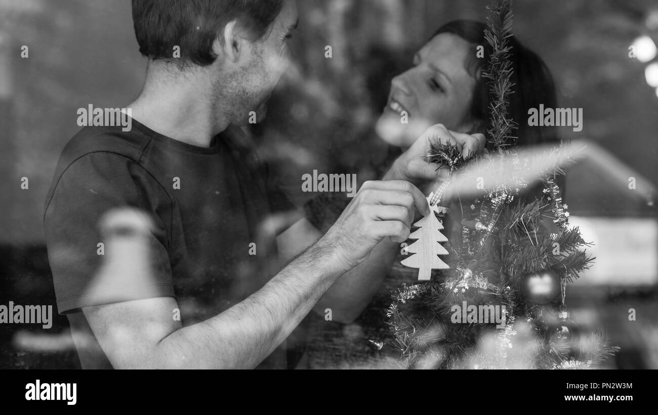 Graustufen Bild eines liebenden jungen Paar schmücken Weihnachtsbaum im Innenbereich. Blick durch ein Fenster mit der winterlichen Natur in einem Glas widerspiegelt. Stockfoto