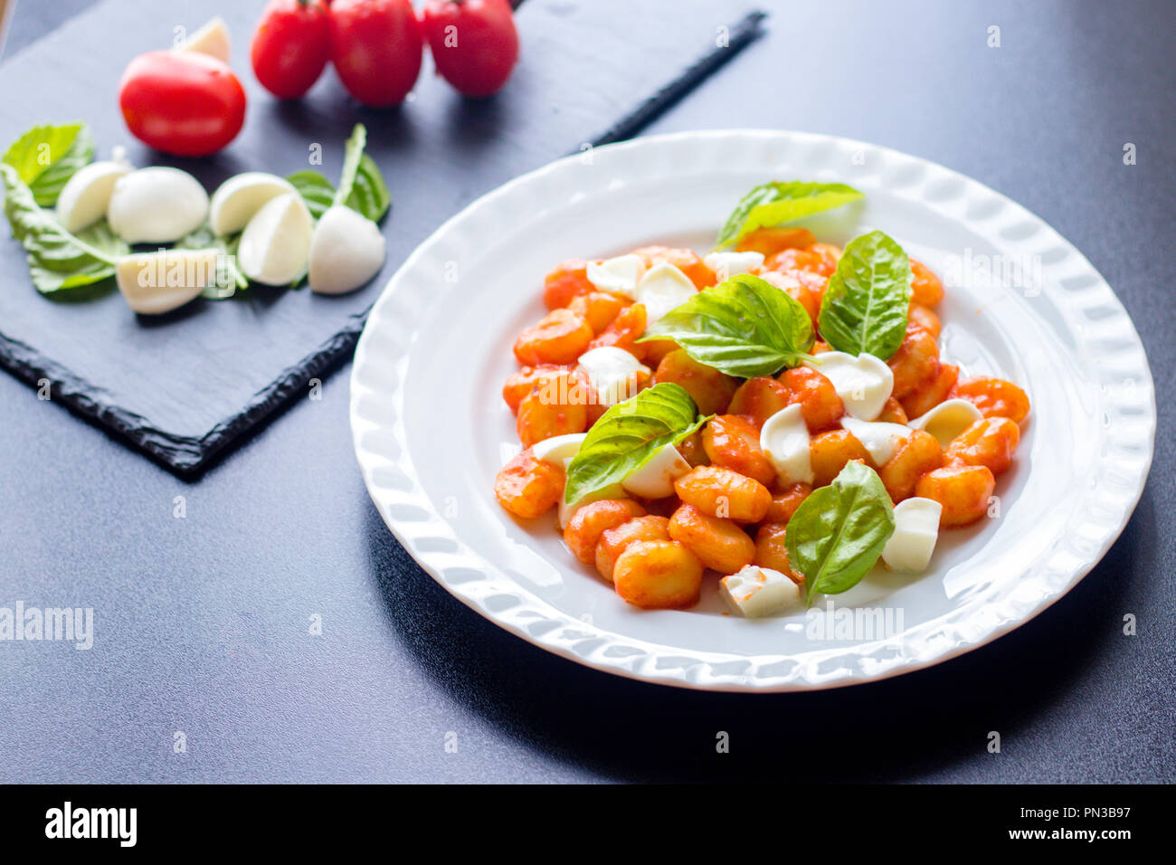 Gnocchi alla Sorrentina in Tomatensauce mit frischem Basilikum und Mozzarella in Scheiben geschnittene Kugeln auf einem weißen Teller mit Zutaten auf dem Schwarzen Brett bac serviert. Stockfoto