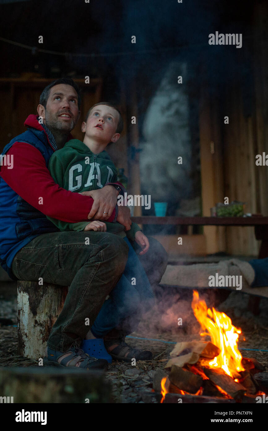 Vater und Sohn sitzen rund um ein Lagerfeuer in der Nacht lachen, reden und Blick auf die Sterne. Stockfoto