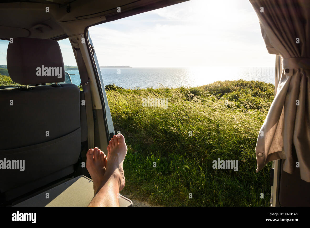 First Person Ansicht eines barfuß Mann entspannen in einem Wohnmobil und genießen den Blick über das Meer bei Sonnenuntergang durch die geöffnete Schiebetür. Stockfoto