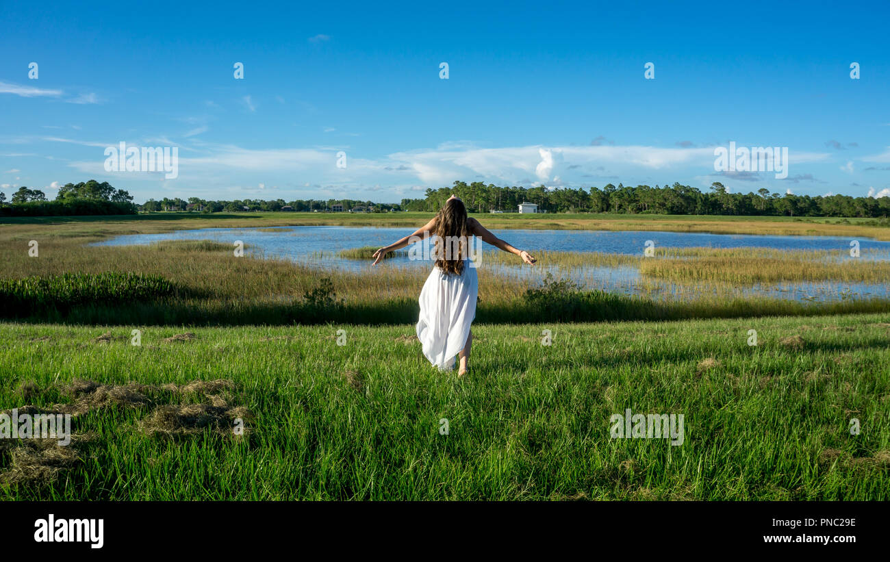 Rückseite der Brünette junge Frau Braut mit langen Locken tragen weiße Walking ausgefahrenen Armen umfassende eart und Sky in einer Landschaft neben einem See durin Stockfoto