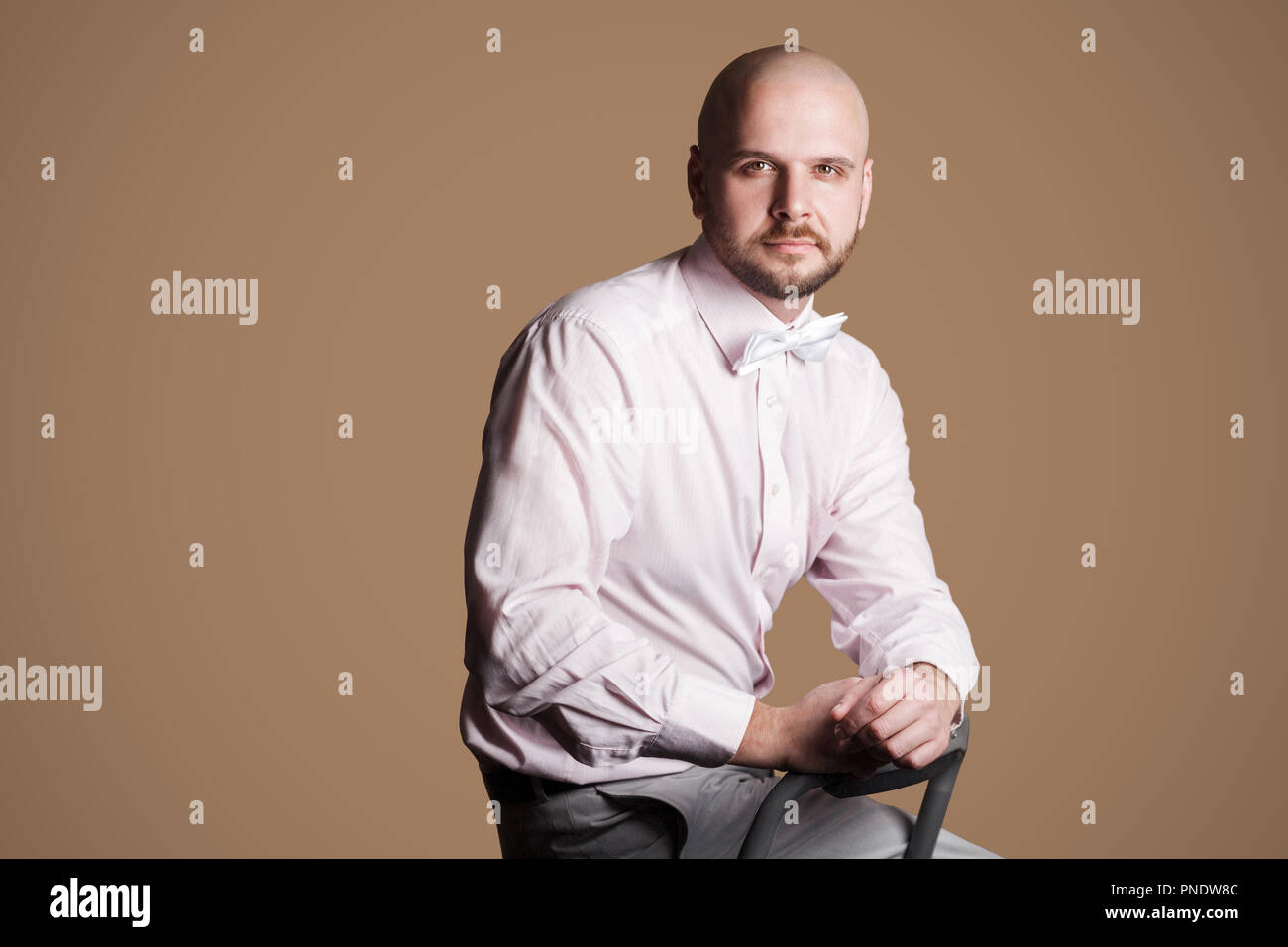 Portrait von erfolgreichen aussehenden Bärtigen glatzköpfige Mann in rosa Hemd und weißer Bogen, sitzt auf einem Stuhl und Blick auf Kamera mit zuversichtlich. indoor Stockfoto