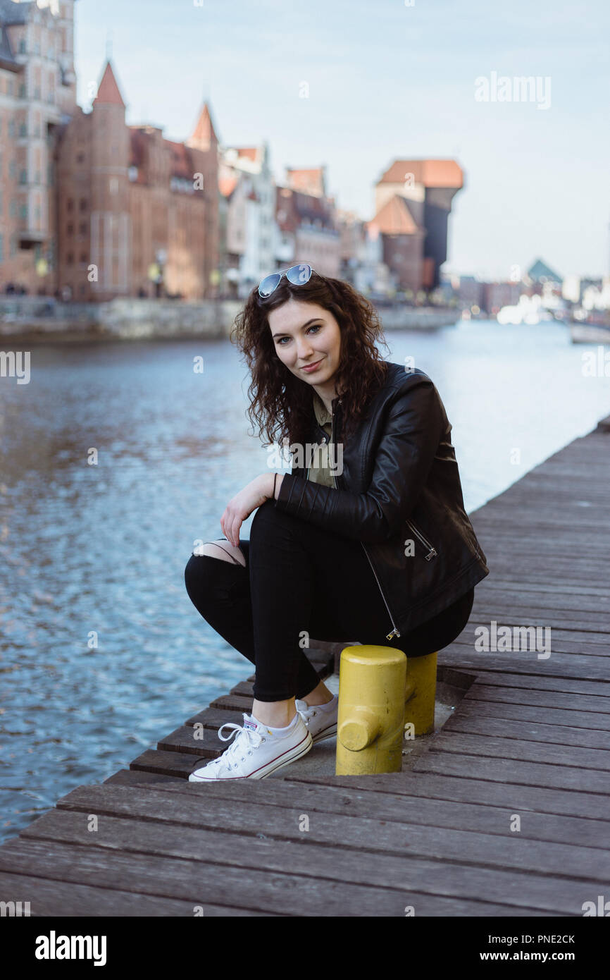 Junge Frau in eine Lederjacke gekleidet sitzt durch den Fluß Pier in Danzig, Polen, lächelnd. Stockfoto