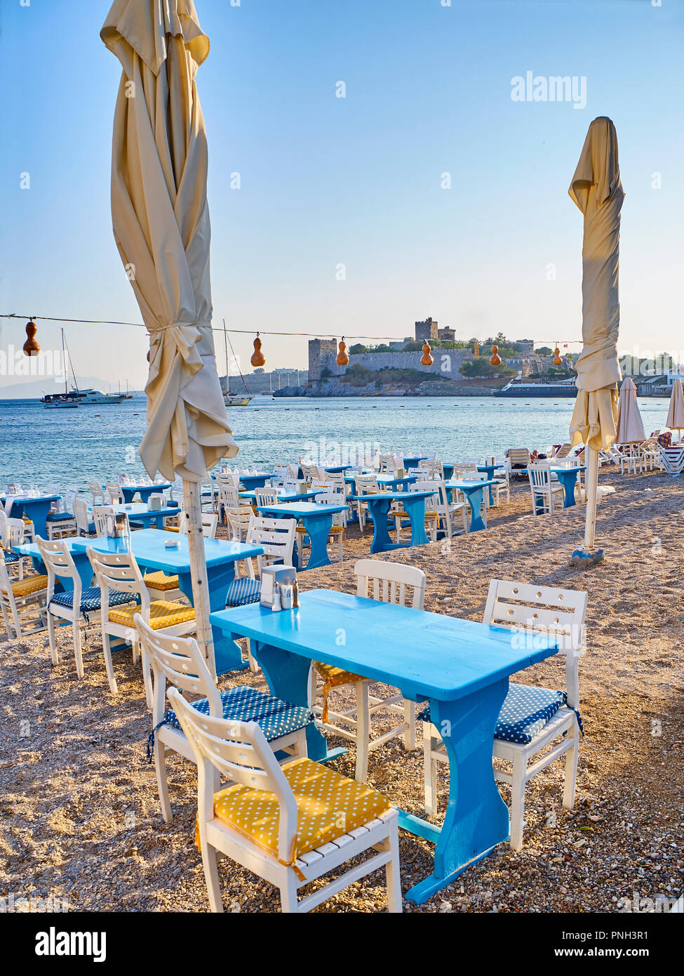 Tische mit Stühlen eines Türkischen Taverne in der Nähe des Meeres in einem Strand der Kumbahce Bucht mit dem Schloss von St. Peter im Hintergrund. Bodrum. Die Türkei. Stockfoto