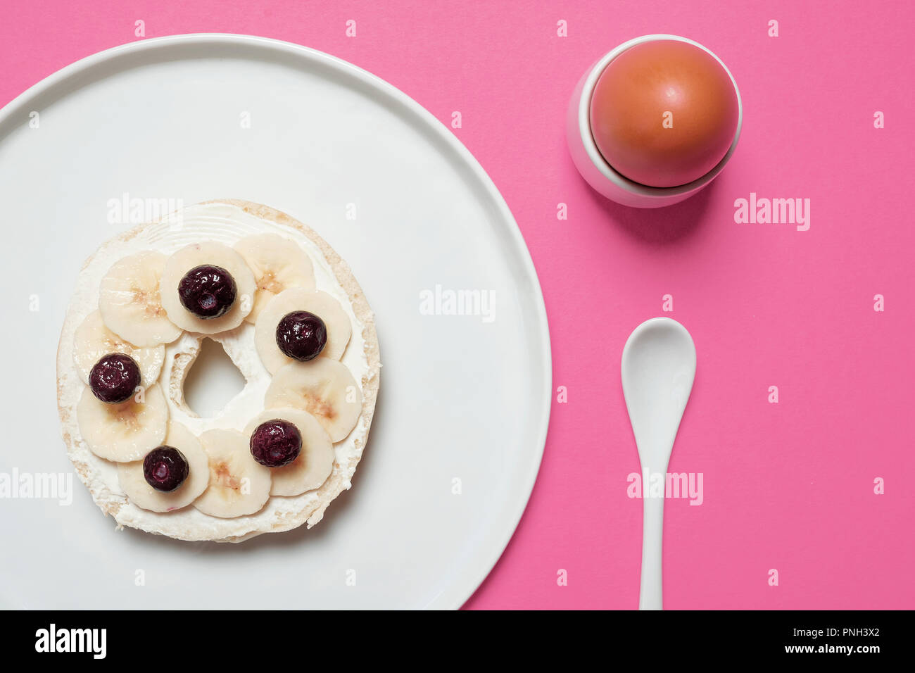 Die Hälfte Bagel auf weiße Platte mit Frischkäse, Blaubeeren, und in Scheiben geschnittene Banane mit einem hart gekochten Ei und weiß Löffel auf einem rosa Hintergrund Stockfoto