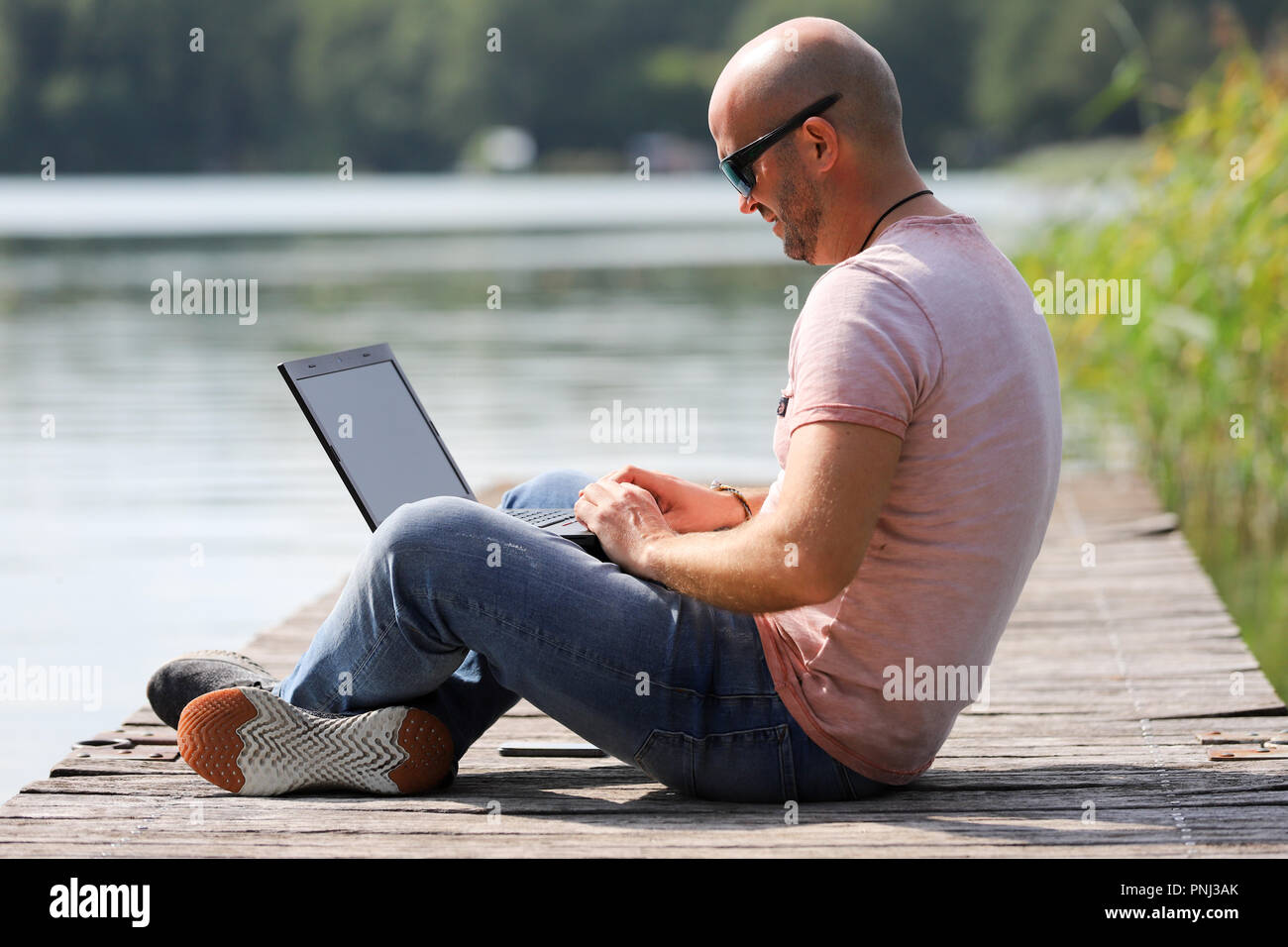 Der 41-jährige sportlich Mann arbeitet in Brandenburg in der Nähe von einem See mit Smartphone und Laptop. Stockfoto