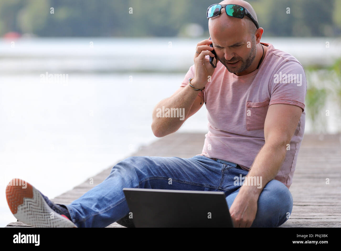 Der 41-jährige sportlich Mann arbeitet in Brandenburg in der Nähe von einem See mit Smartphone und Laptop. Stockfoto