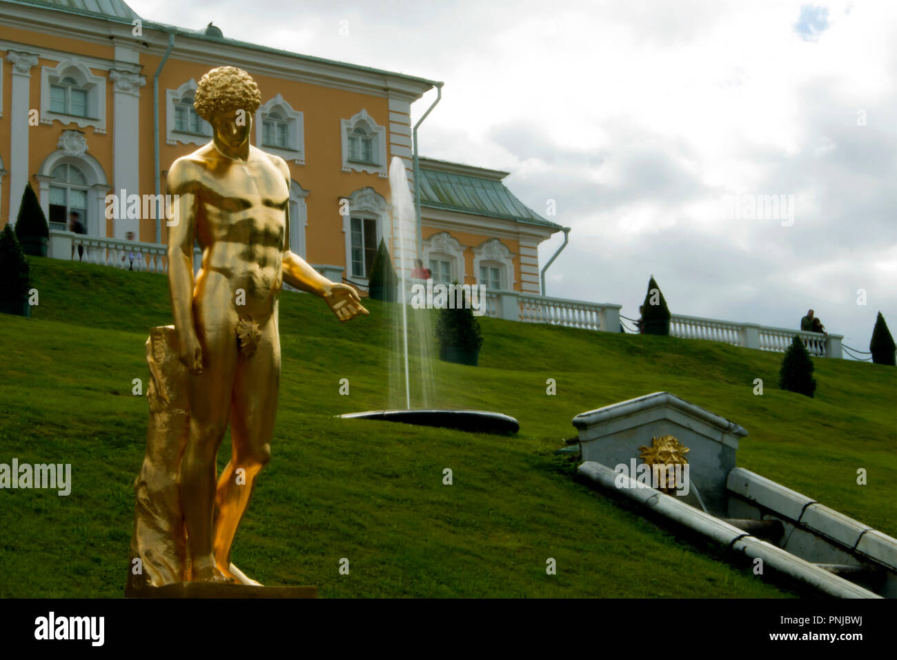 Vergoldete Statue des Kapitolinischen Antinous gegen grasigen Hang mit Brunnen und großen Schloss Peterhof in St. Petersburg, Russland, 18. Jahrhundert Sehenswürdigkeiten Stockfoto