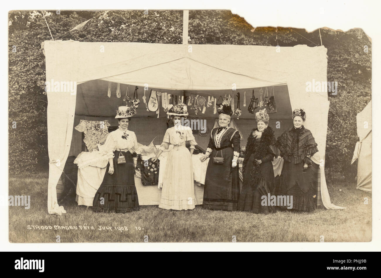 Original-Fotopostkarte aus der Zeit Edwardians zur Sommerzeit von Damen im Strood Garden Fete, die für ein Foto von einem Stall posiert, der handgenähte Geschenke und handgefertigte Handarbeiten verkauft - wunderbare Bilder von Hüten, Frauen unterschiedlichen Alters, atemberaubende Outfits. Postkarte vom Juli 1908, Strood, Kent, Großbritannien Stockfoto