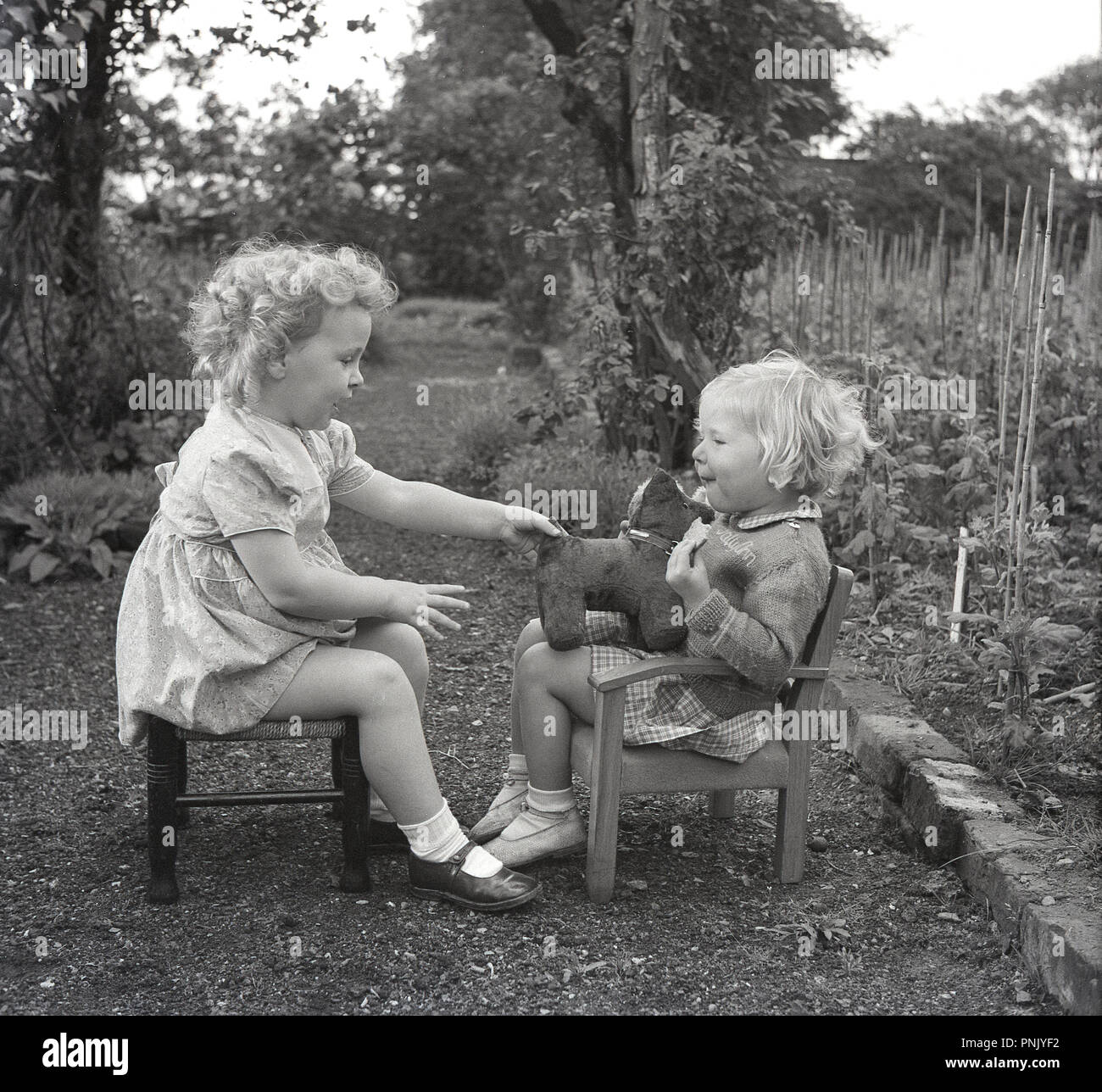 1950, historische, zwei kleine Kinder sitzen im Freien einander gegenüber zu wenig Sitzplätze auf einem Gartenweg Spaß mit einem kleinen Kuscheltier Hund, England, UK. Stockfoto