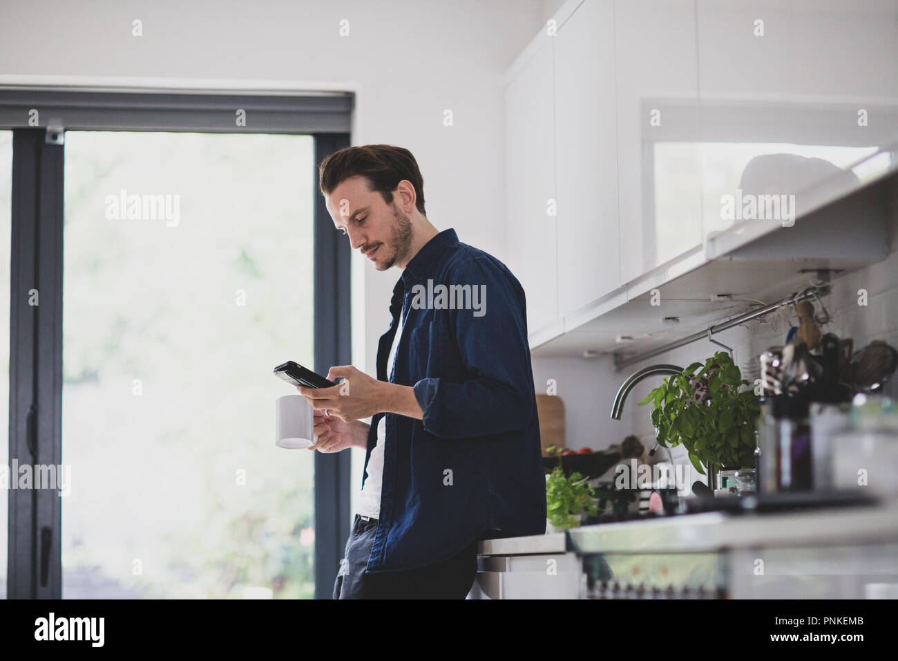 Erwachsene männliche Kontrolle Smartphone in Küche mit Becher Kaffee Stockfoto
