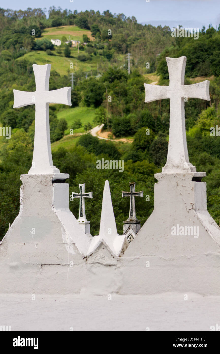 Asturien, Spanien. Weißer Stein Kreuze auf dem Friedhof mit einem Bergdorf im Hintergrund. Stockfoto