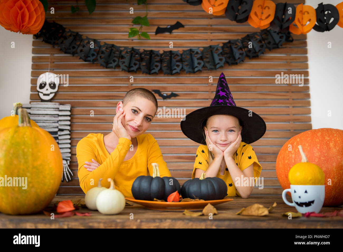 Süße kleine Mädchen und ihre Mutter, ein Krebspatient, hinter einem Tisch in Halloween Thema eingerichtete Zimmer sitzen, in die Kamera schauen und lächeln. Familie Hal Stockfoto