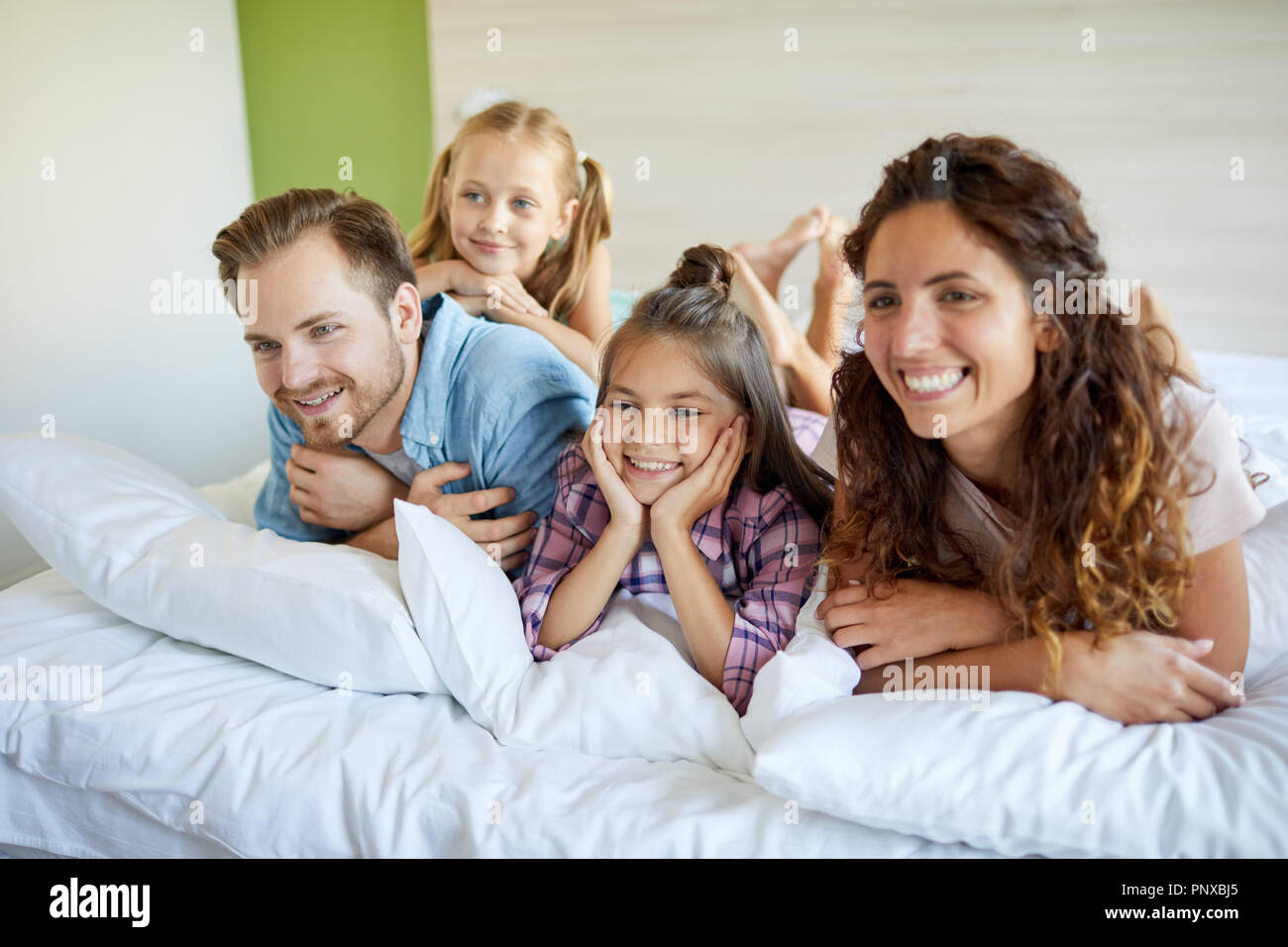 Junge fröhliche Familie von Eltern und zwei kleinen Töchtern liegend auf dem Bett vor dem Fernseher in der Freizeit gesetzt Stockfoto