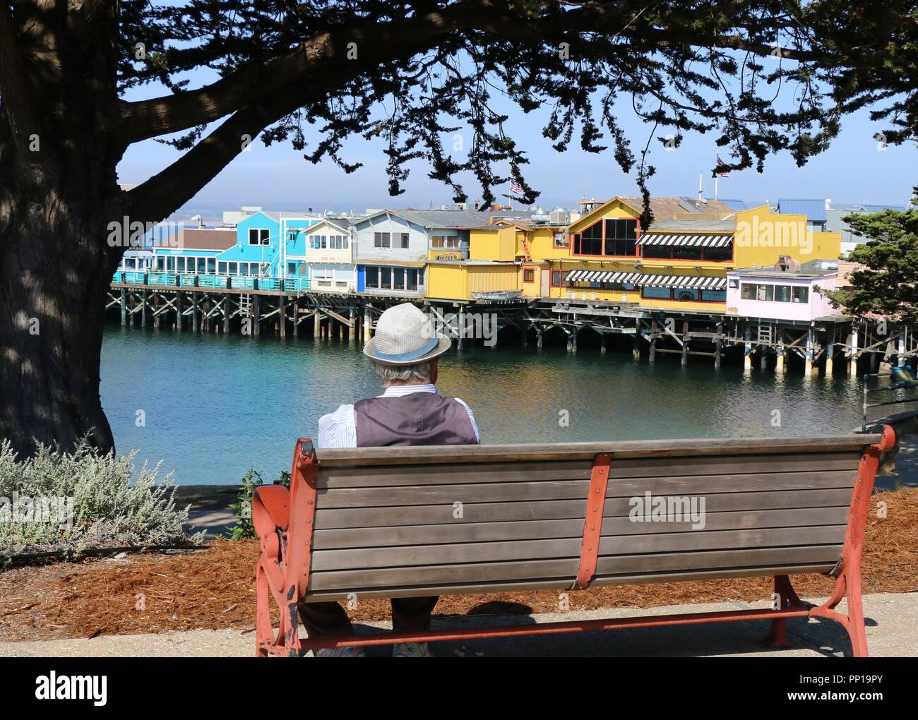 Ein älterer Mann mit einem Adretten Hut sitzt auf einer Bank mit Blick auf Fisherman's Wharf, Monterey, CA. Stockfoto