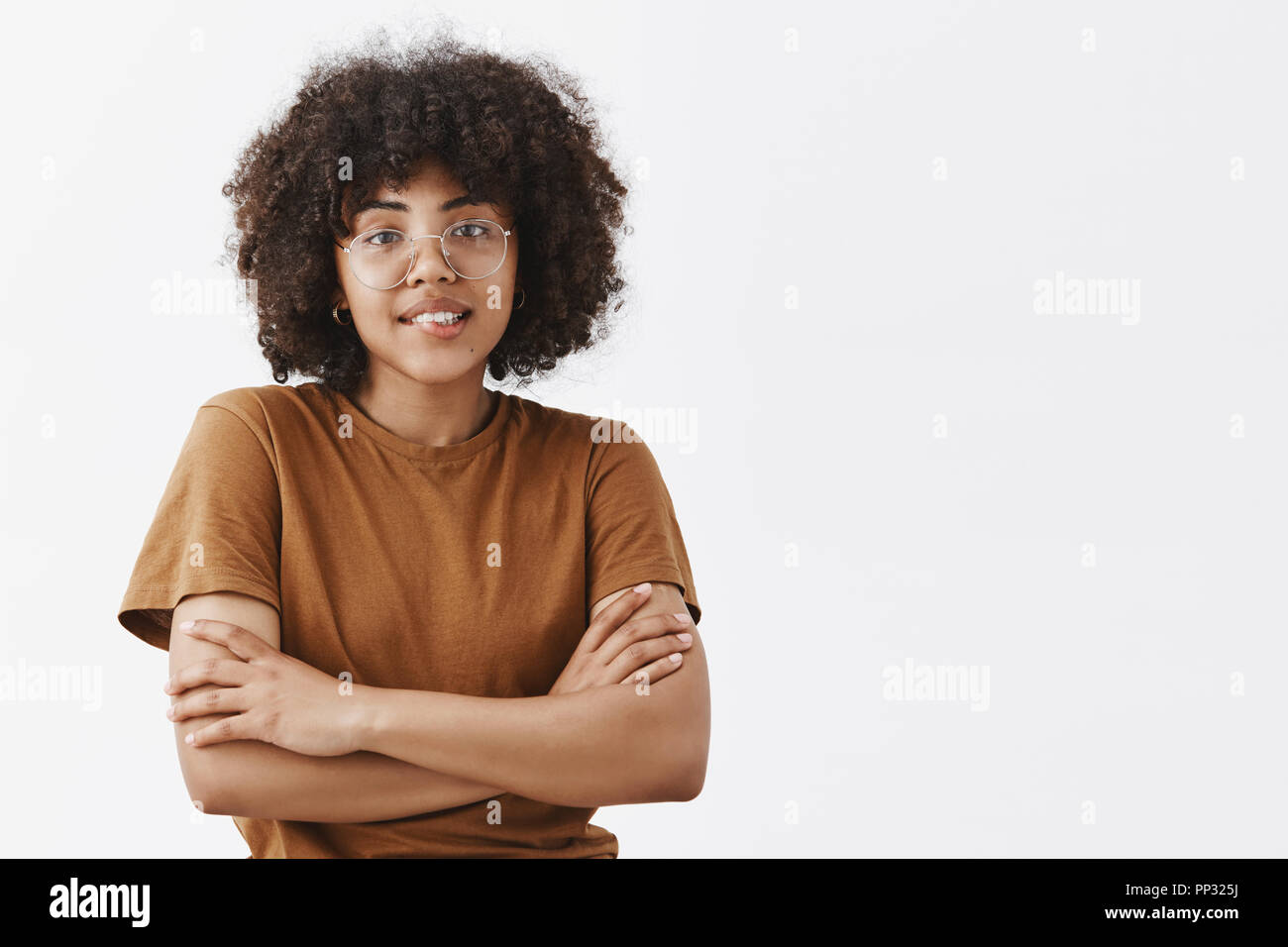 Taille-up Shot schüchtern und albern dunkelhäutigen Jugendlichen weiblichen in transparenten Gläser und trendigen Braunes t-shirt Frieren Kreuzung Hände auf Brust und lächelnd mit freundlich und höflich Ausdruck Stockfoto