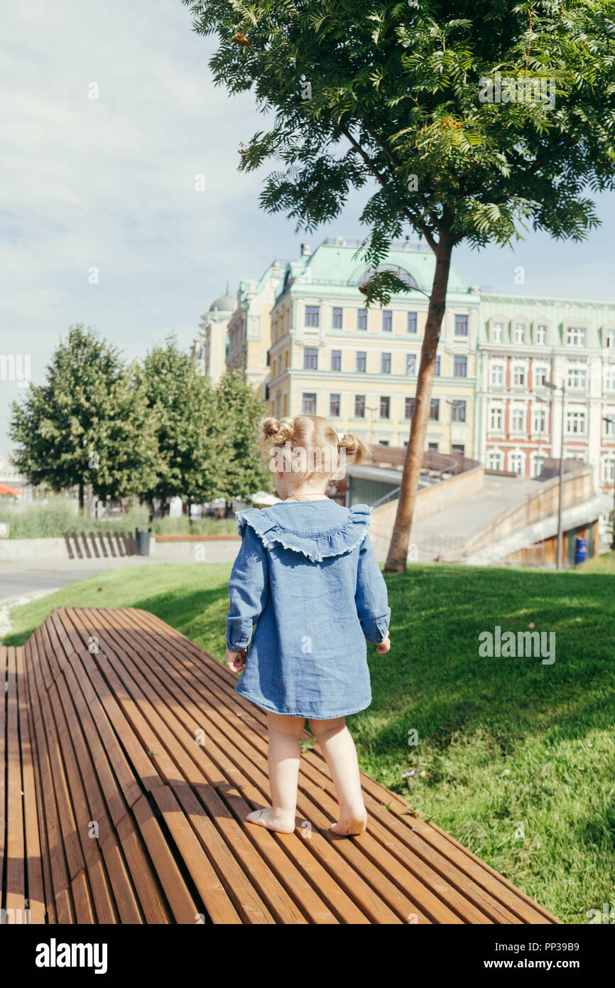 Das kleine Mädchen in blauen Jeans Kleid barfuß laufen allein Auf einer Holzbank in einem Stadtpark in der Zentrum von Moskau an einem warmen sonnigen Tag Stockfoto