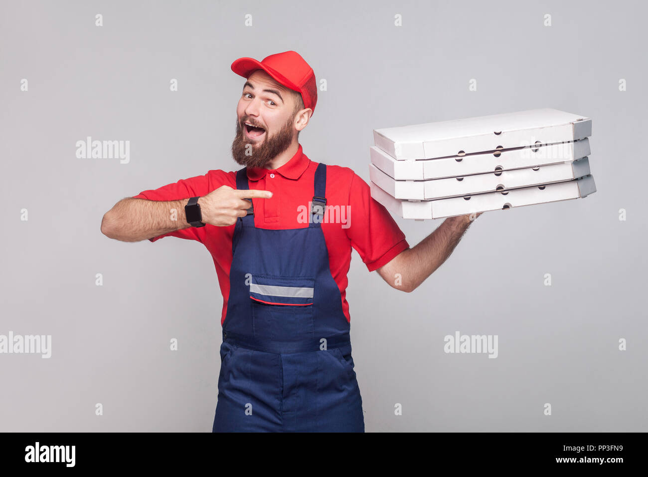 Junge glücklich Lieferung Mann mit Bart in der blauen Uniform und dem roten T-Shirt, die hielten und Zeigefinger aus Karton Pizzakartons auf Grau ba zu Stapeln Stockfoto