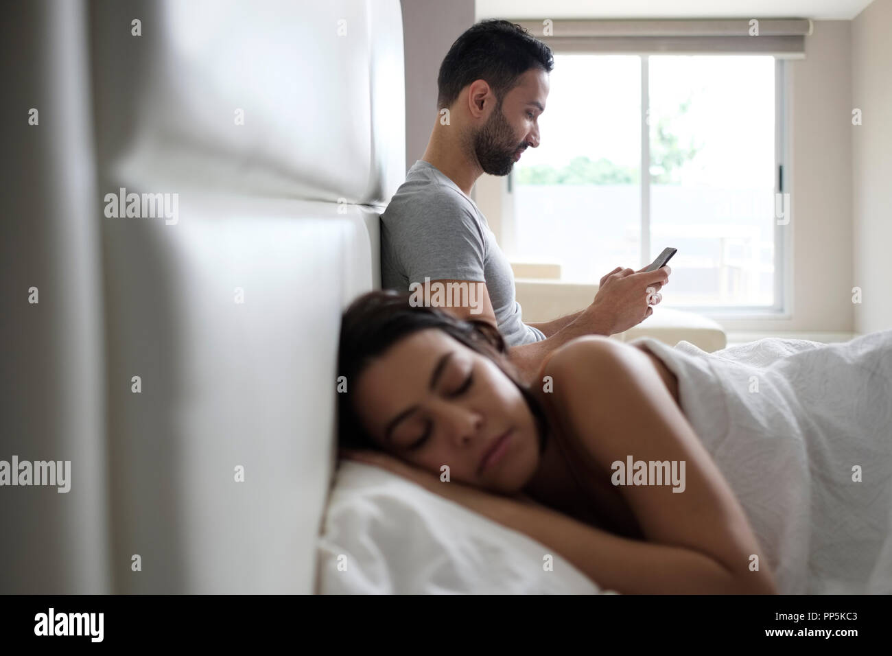 Jungen Latino Mann und Frau im Bett zu Hause. Spanischer Mann texting mit Smartphone und Senden der Nachricht zu seinem Geliebten, während seine Frau noch schläft Stockfoto